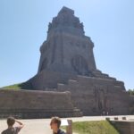 Außenaufnahme seitlich vom Völkerschlachtdenkmal. Riesiges monumentales, steinernes Gebäude, das sich verjüngend von einem breiten Sockel aufragt.