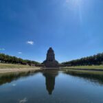 Blick über eine Wasserfläche auf das steilaufragende Völkerschlachtdenkmal vor blauem Himmel in hellem Sonnenlicht.