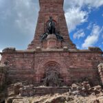 Das Kyffhäuserdenkmal in Gänze frontral fotografiert. Unten sitzt eine große Statue des Barbarossa, ein alter Mann mit langem Bart, darüber trohnt vor einer Stele aus rotem Stein ein Reiterstandbild Kaiser Wilhelm I.