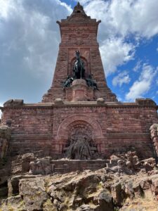 Das Kyffhäuserdenkmal in Gänze frontal fotografiert. Unten sitzt eine große Statue des Barbarossa, ein alter Mann mit langem Bart, darüber trohnt vor einer Stele aus rotem Stein ein Reiterstandbild Kaiser Wilhelm I.
