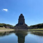 Blick über eine Wasserfläche auf das steilaufragende Völkerschlachtdenkmal vor blauem Himmel in hellem Sonnenlicht.