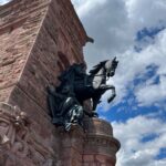 Das Reiterstandbild Wilhelm I. seitlich von unten fotografiert. Im Hintergrund der rote Stein des Denkmals und blauer Himmel mit weißen Fetzenwolken.