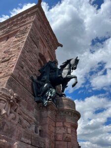 Das Reiterstandbild Wilhelm I. seitlich von unten fotografiert. Im Hintergrund der rote Stein des Denkmals und blauer Himmel mit weißen Fetzenwolken.
